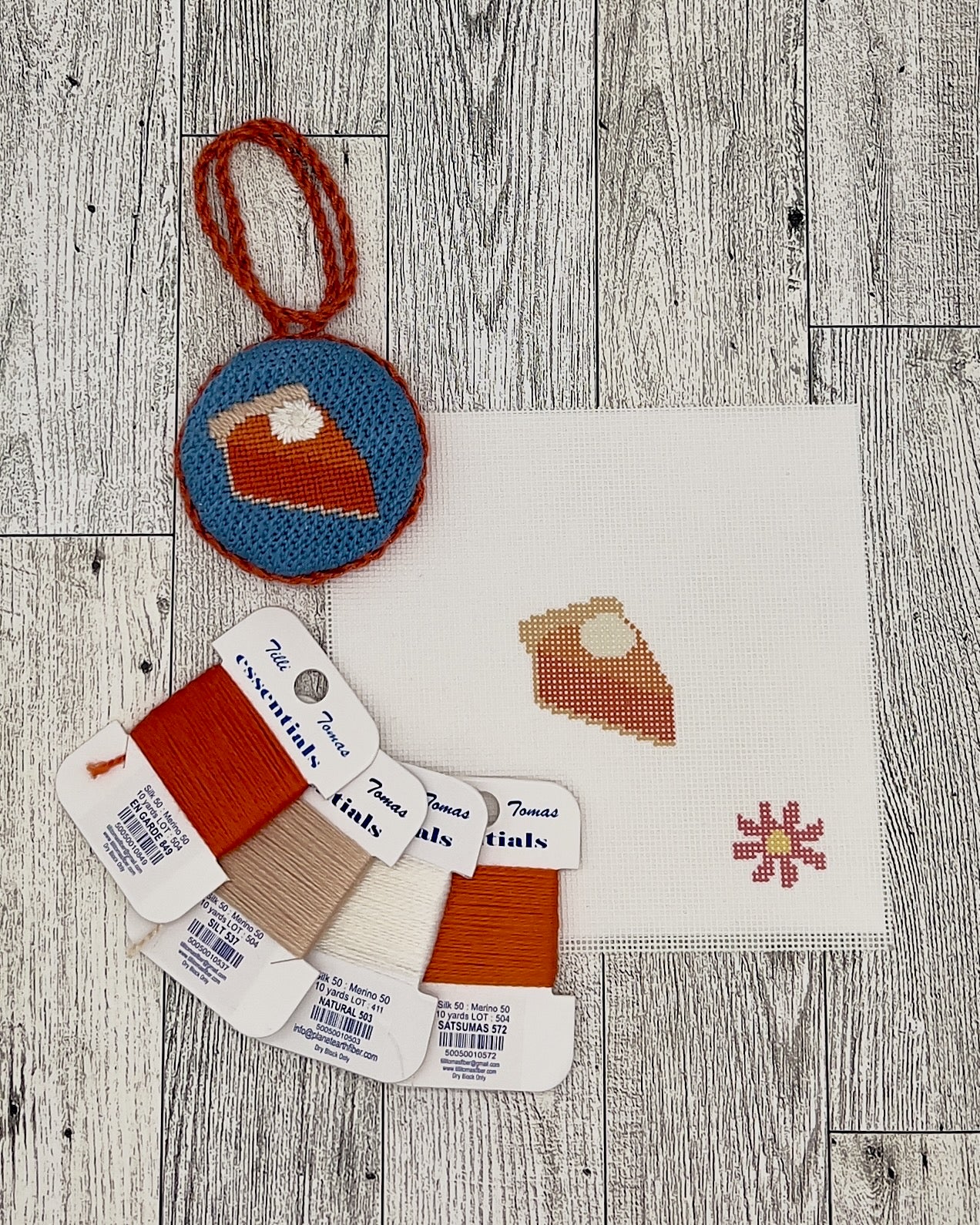 various craft supplies, including a needlepoint kit, thread, and a small embroidery piece, all arranged on a wooden surface.