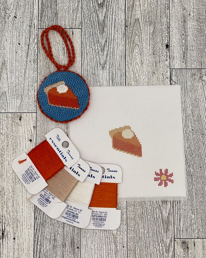 various craft supplies, including a needlepoint kit, thread, and a small embroidery piece, all arranged on a wooden surface.